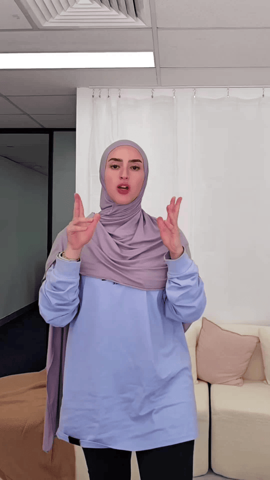 A person wearing a light purple hijab and light blue shirt gestures with both hands while standing indoors.