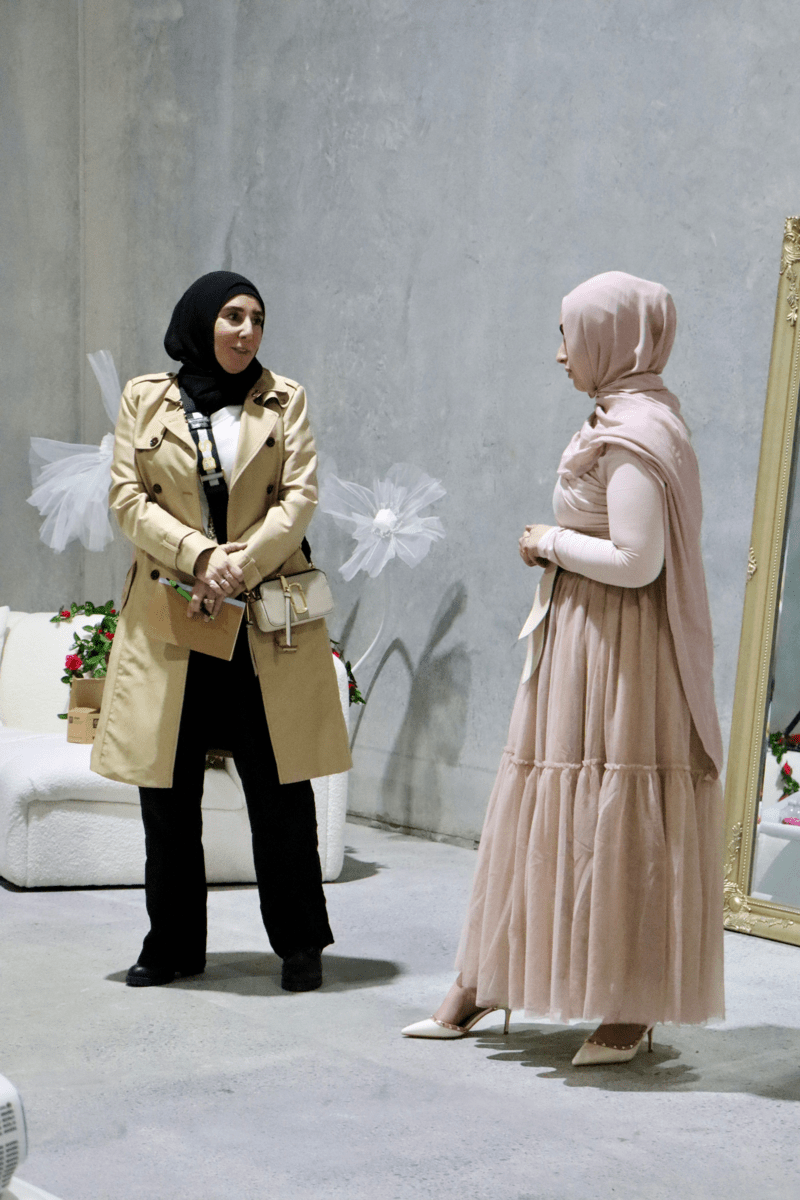 Two individuals engaged in a conversation, one wearing a beige coat and the other in a pink dress, set against a simple backdrop.