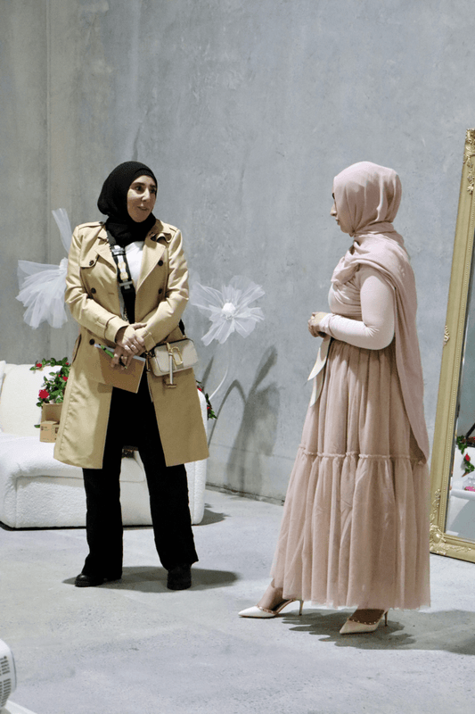 Two individuals engaged in a conversation, one wearing a beige coat and the other in a pink dress, set against a simple backdrop.