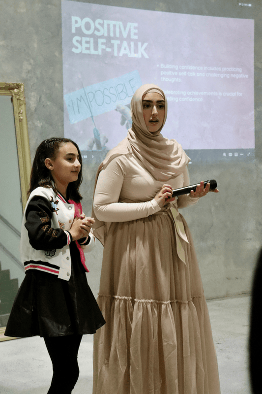A photo showing two individuals, possibly a counselor and a child client, presenting in front of a slide titled 'POSITIVE SELF-TALK'. The child is wearing a black jacket and the counselor is in a beige dress with a hijab.