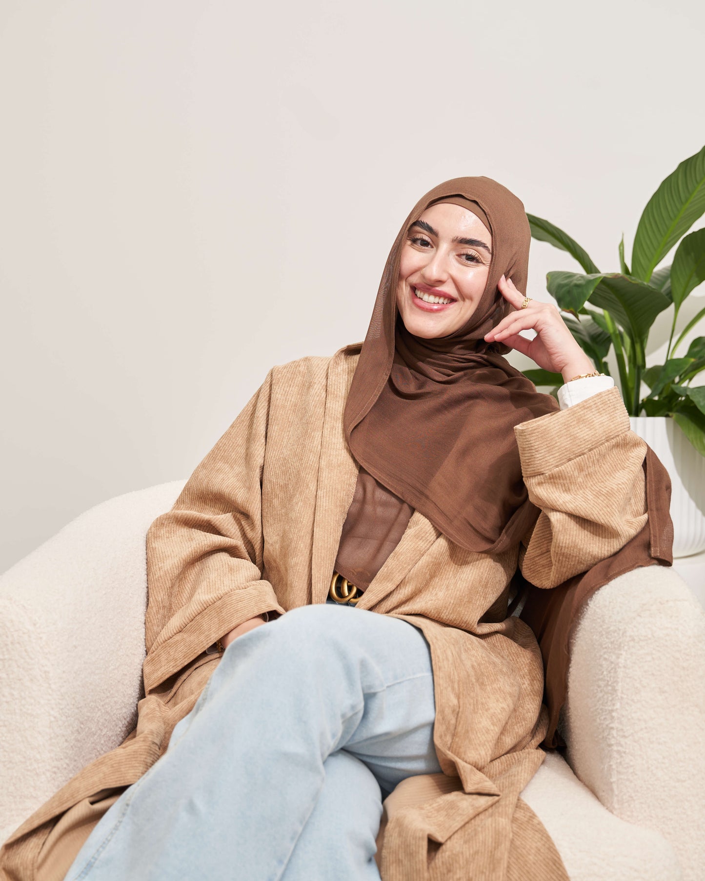 Woman wearing a brown hijab and beige coat sitting on a white chair with a plant in the background.